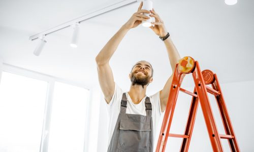 Repairman or professional electrician in workwear installing light spots, standing on the ladder in the white living room